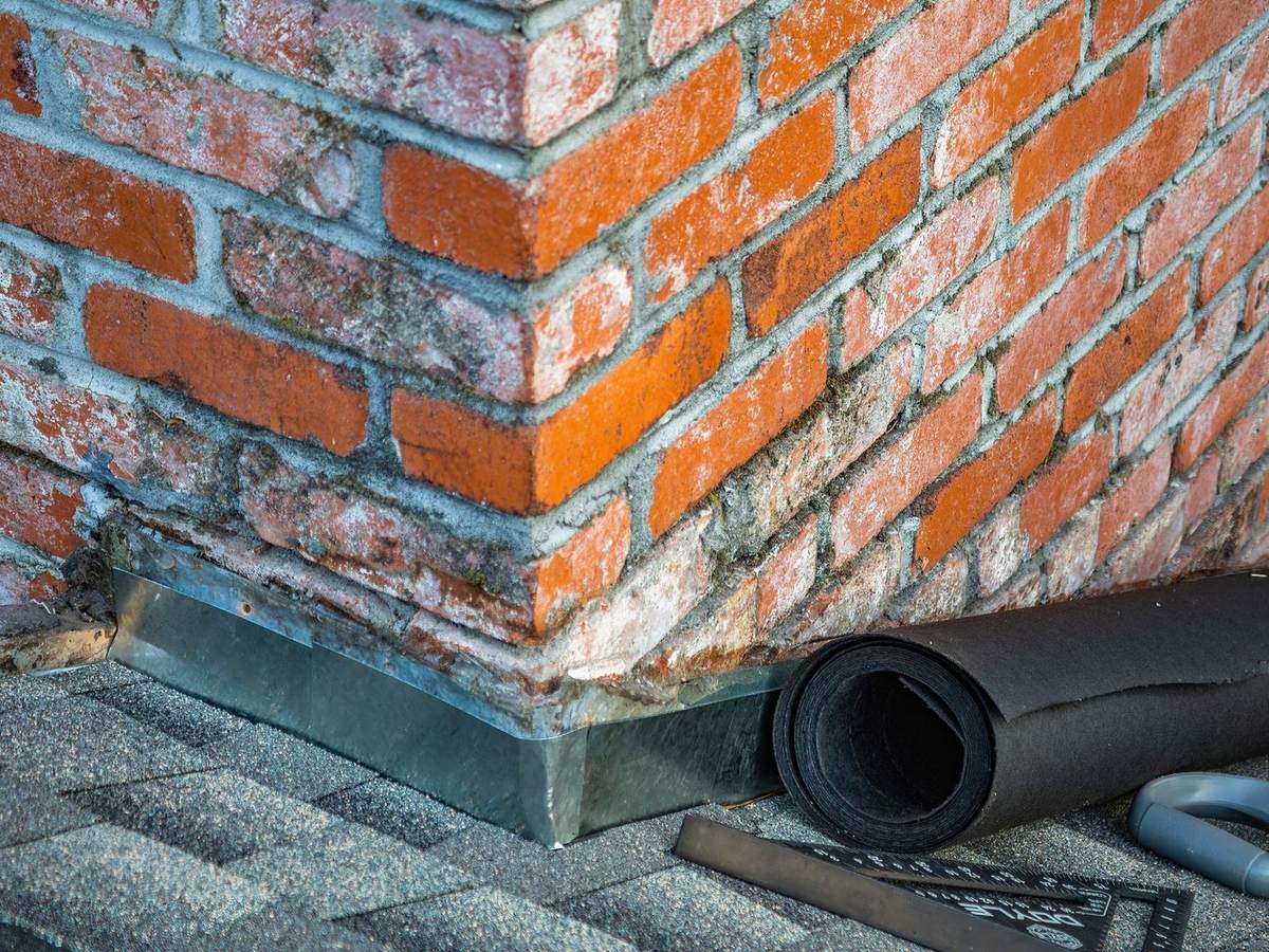 New step flashing at the base of a brick chimney, with a roll of underlayment on the shingles